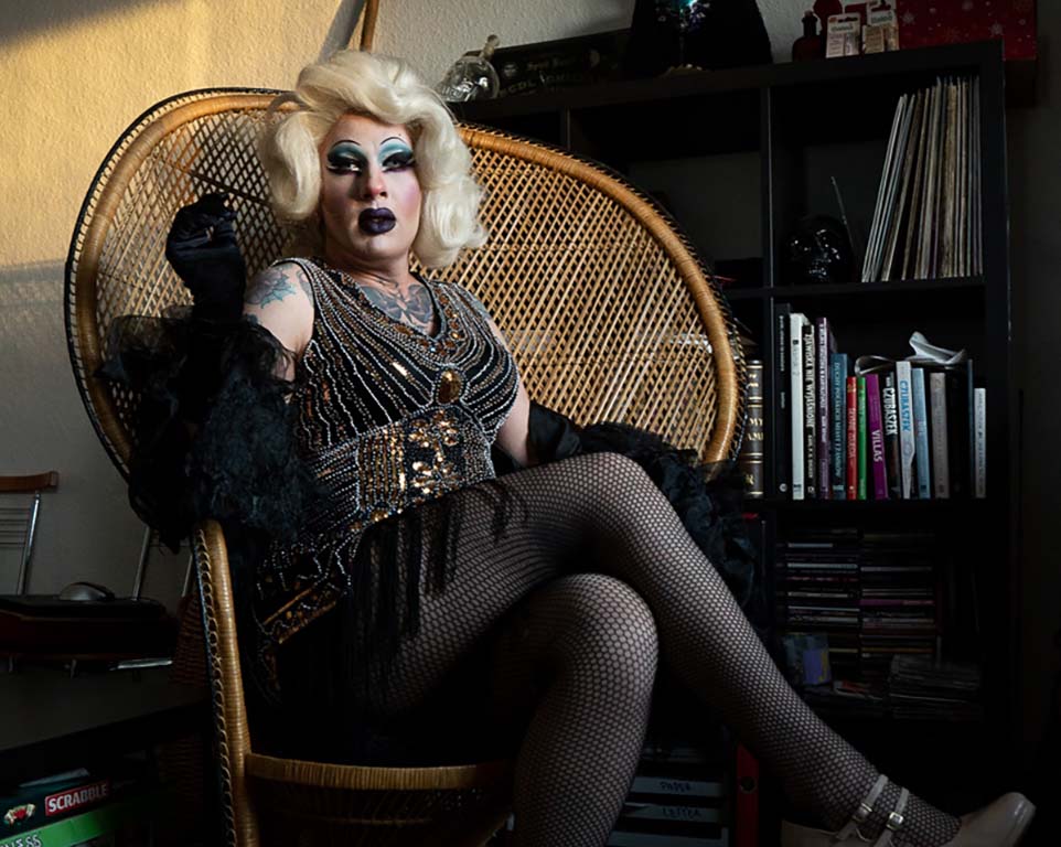 Bild: Drag Queen mit blonder Perücke, blauem Augen Makeup, schwarzen Lippen und langen Wimpern, Tattoo auf der rechten Schulter in einem Korbsessel sitzend das rechte Bein überschlagend, schwarze Netzstrümpfe, glitzernder Body mit Fransen, ein Bücherregal im Hintergrund.