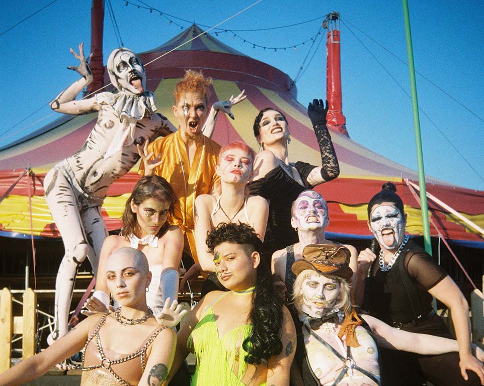Image: Venus Boys 10 young people set up in front of a circus tent all in intense makeup and costumes in wild poses, some of them sticking out their tongues.