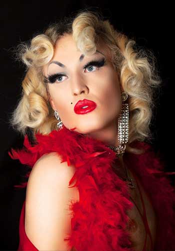 Image: Olive Baldwin looking over her right shoulder, with intense MakeUp, red pouting lips, long glitter earrings, curly blond hair and a red feather boa.