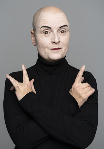 Image: Bridge Markland with shaved head, painted eyebrows and dark lips, arms crossed in front of the body, fingers expressively pointing upwards.