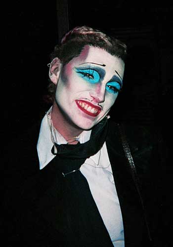 Image : Nancy tilting her head to the left, with intense turquoise eye makeup, painted mustache and eyebrows and grinning red lips.